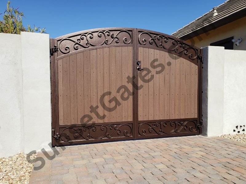 A decorative double gate with metal scrollwork and a wood panel design, set between white concrete walls on a brick driveway, with a house roof visible on the right and foliage on the left under a clear blue sky.