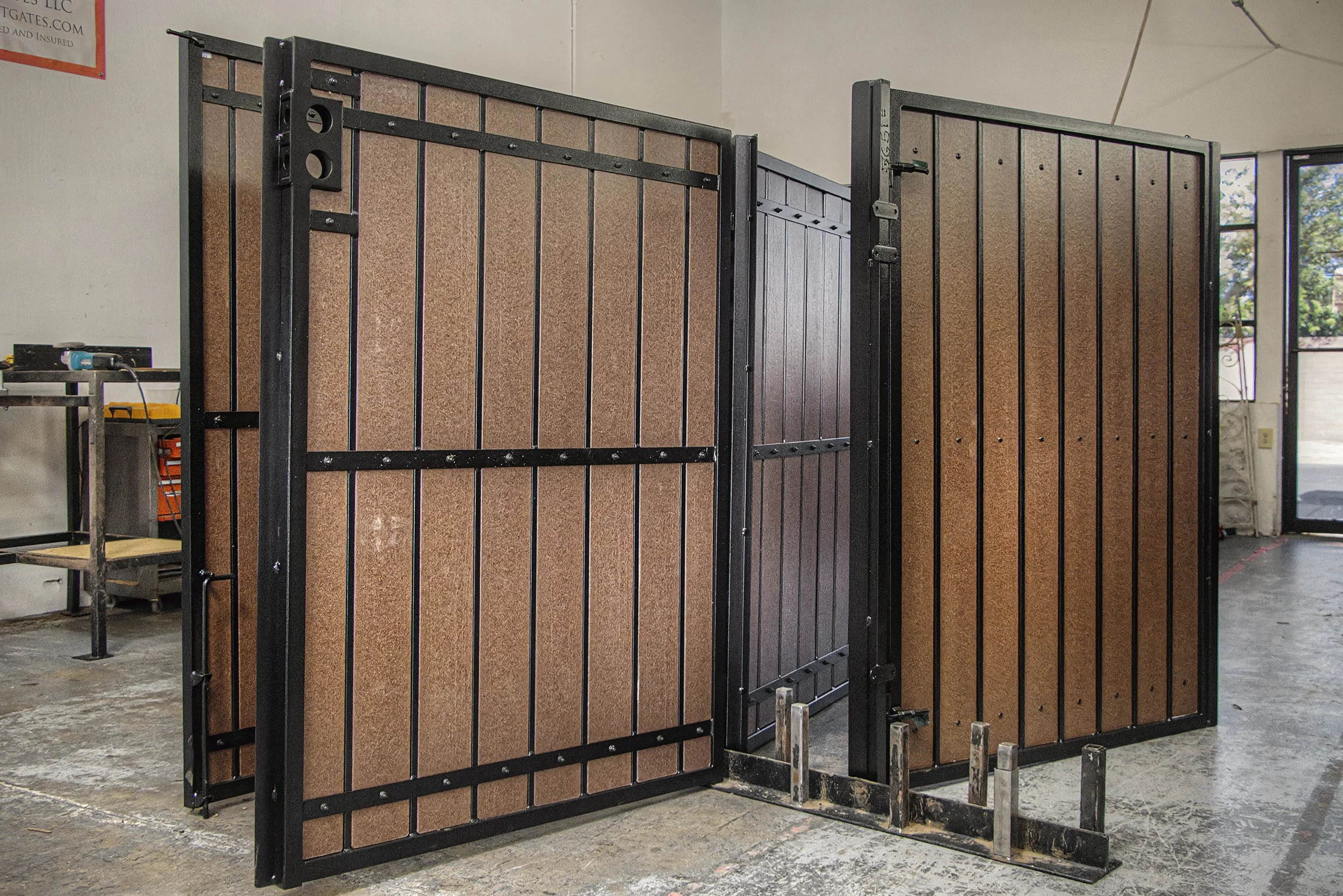 Two large metal and wood fences standing upright on a concrete floor in a workshop or warehouse, with an open door and a window showing trees outside in the background.