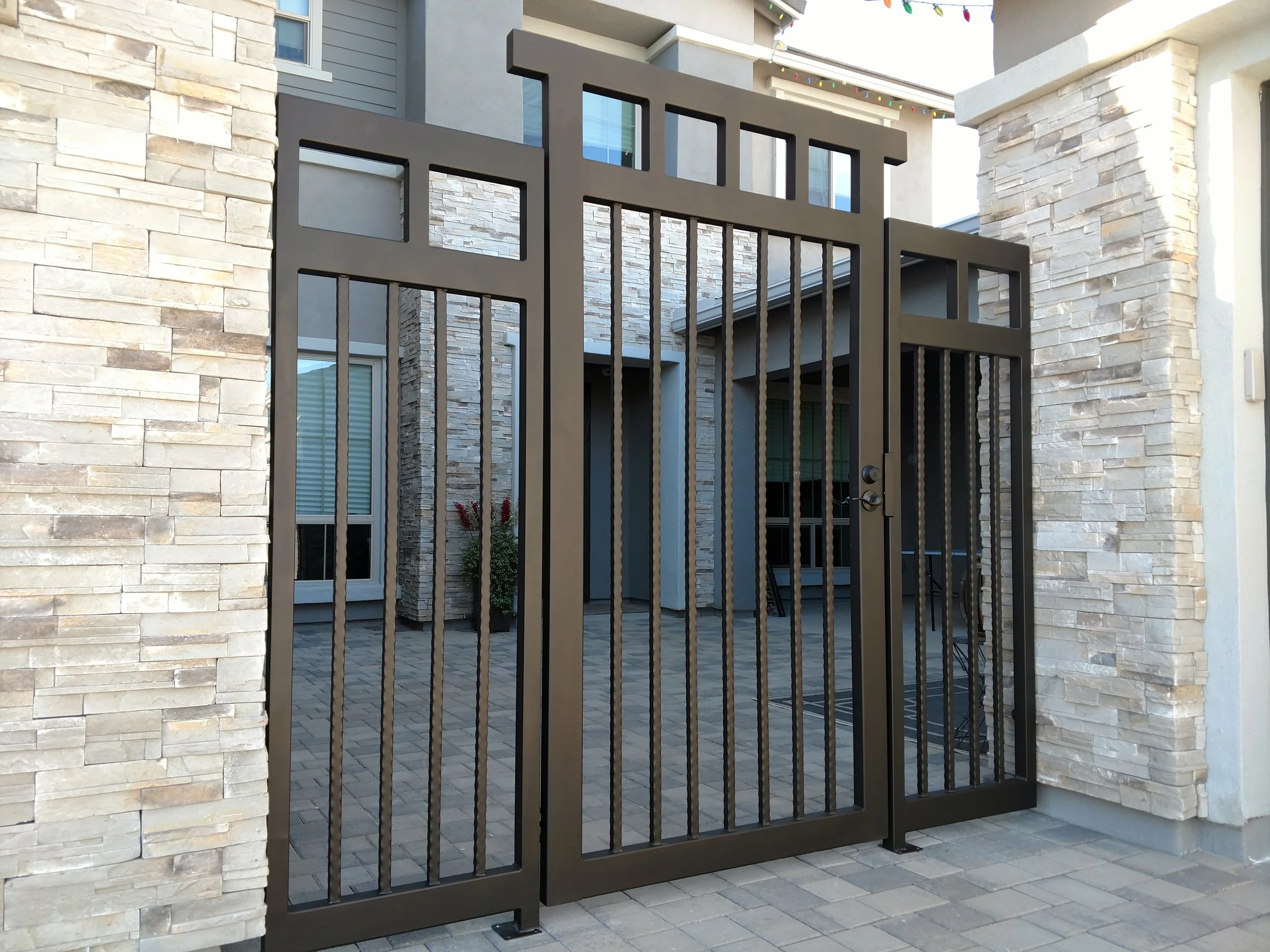 A black metal gate with vertical bars is attached to a stone and stucco wall, leading into a modern residential patio area with pavers and potted plants.