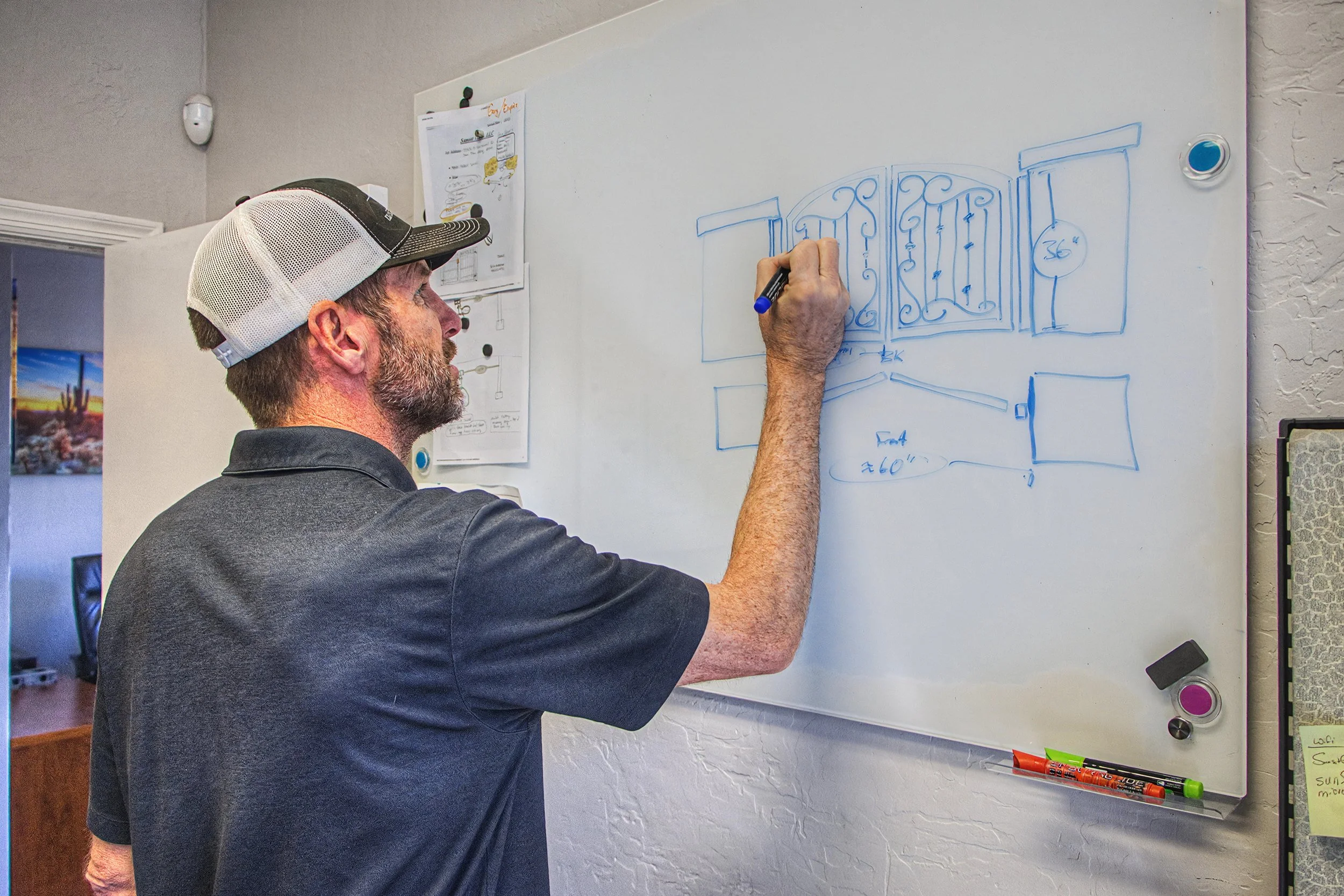 A man with a grey beard and wearing a black and white baseball cap is drawing diagrams on a whiteboard with a blue marker. The whiteboard has several sketches and measurements. The room has a textured wall and a few small objects attached to or near the whiteboard.