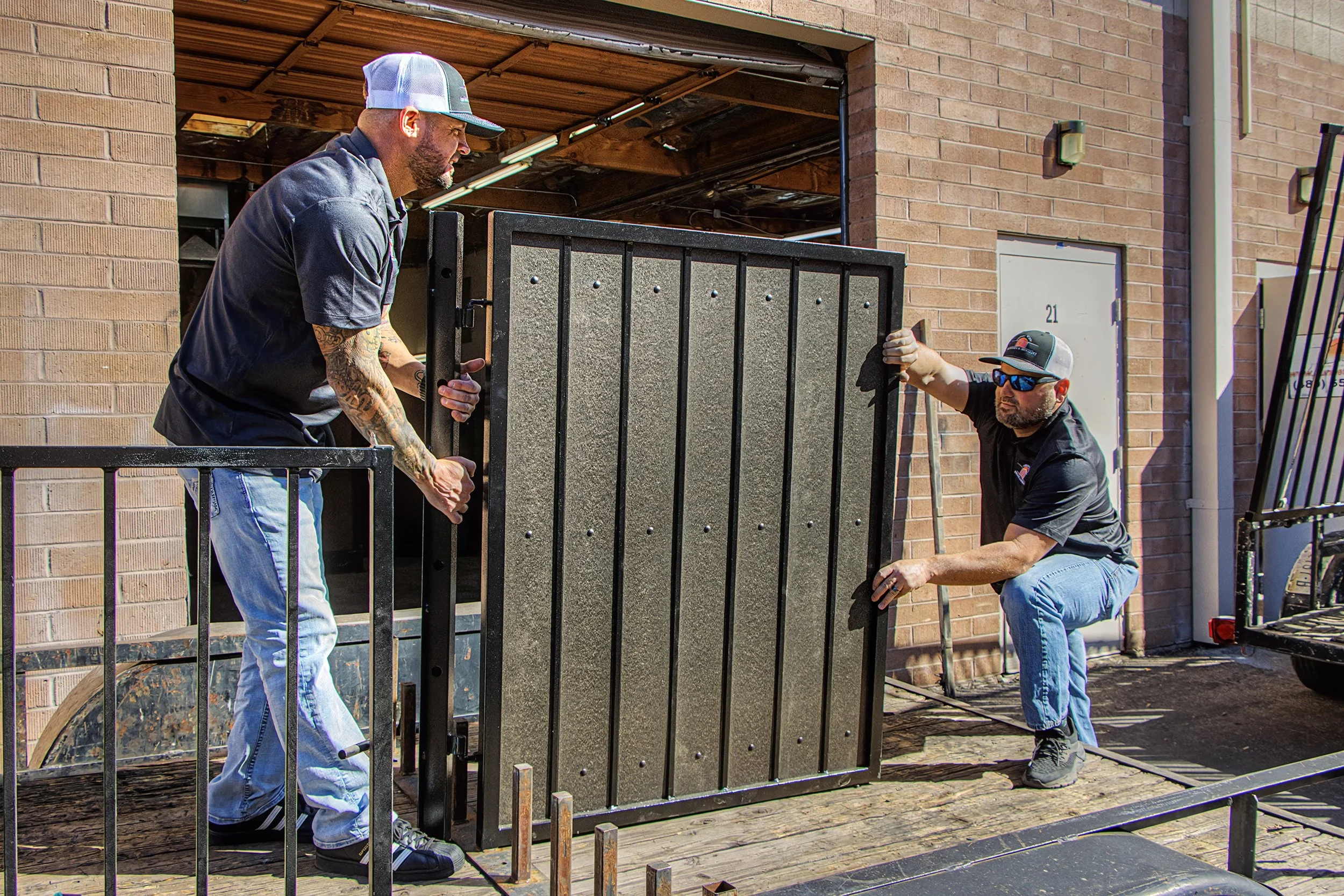 Two men are installing a security gate on a doorway, one is standing on the ground and the other is on a raised platform, working together to hold and position the gate.