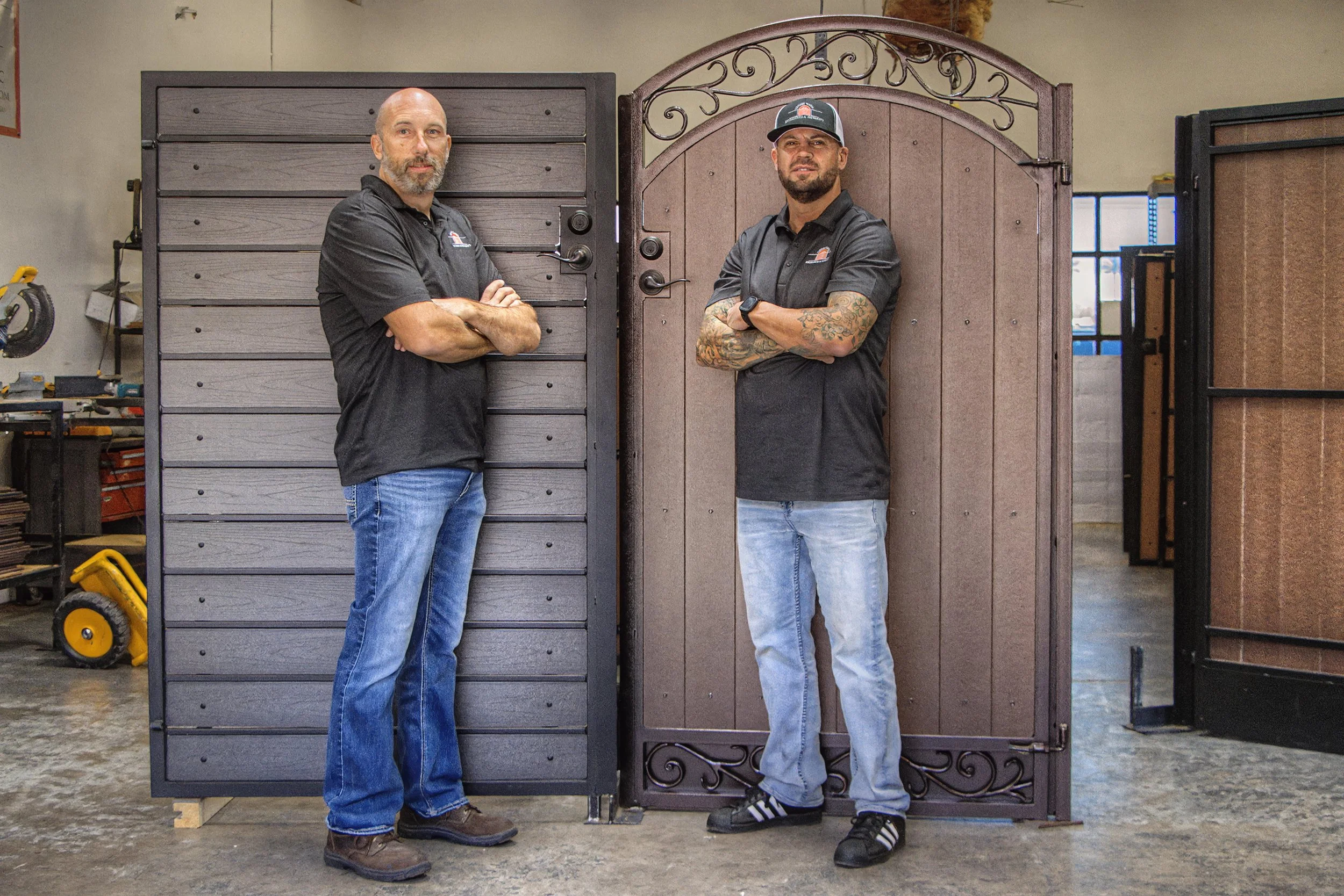 Two men standing with arms crossed in an indoor warehouse, flanked by windowed and wooden large gates or doors. One man has a shaved head and gray beard, wearing a black polo shirt and jeans. The other man has tattoos on his arms, a beard, and is wearing a black shirt, light jeans, and a cap.