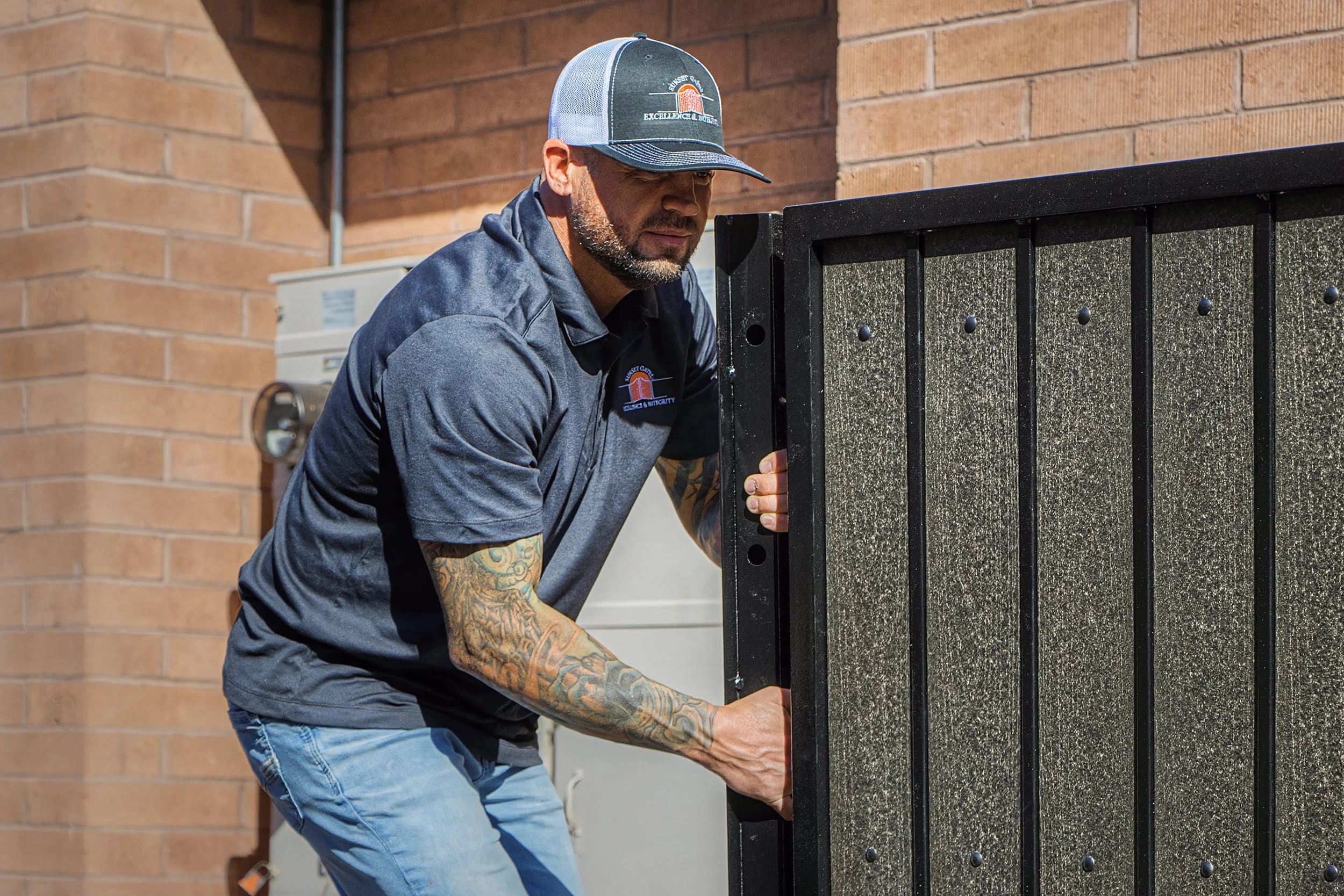 A man with tattoos on his arm and a beard, wearing a trucker hat and a gray jacket, is installing or repairing a black wall-mounted fire sprinkler system outside a brick building.