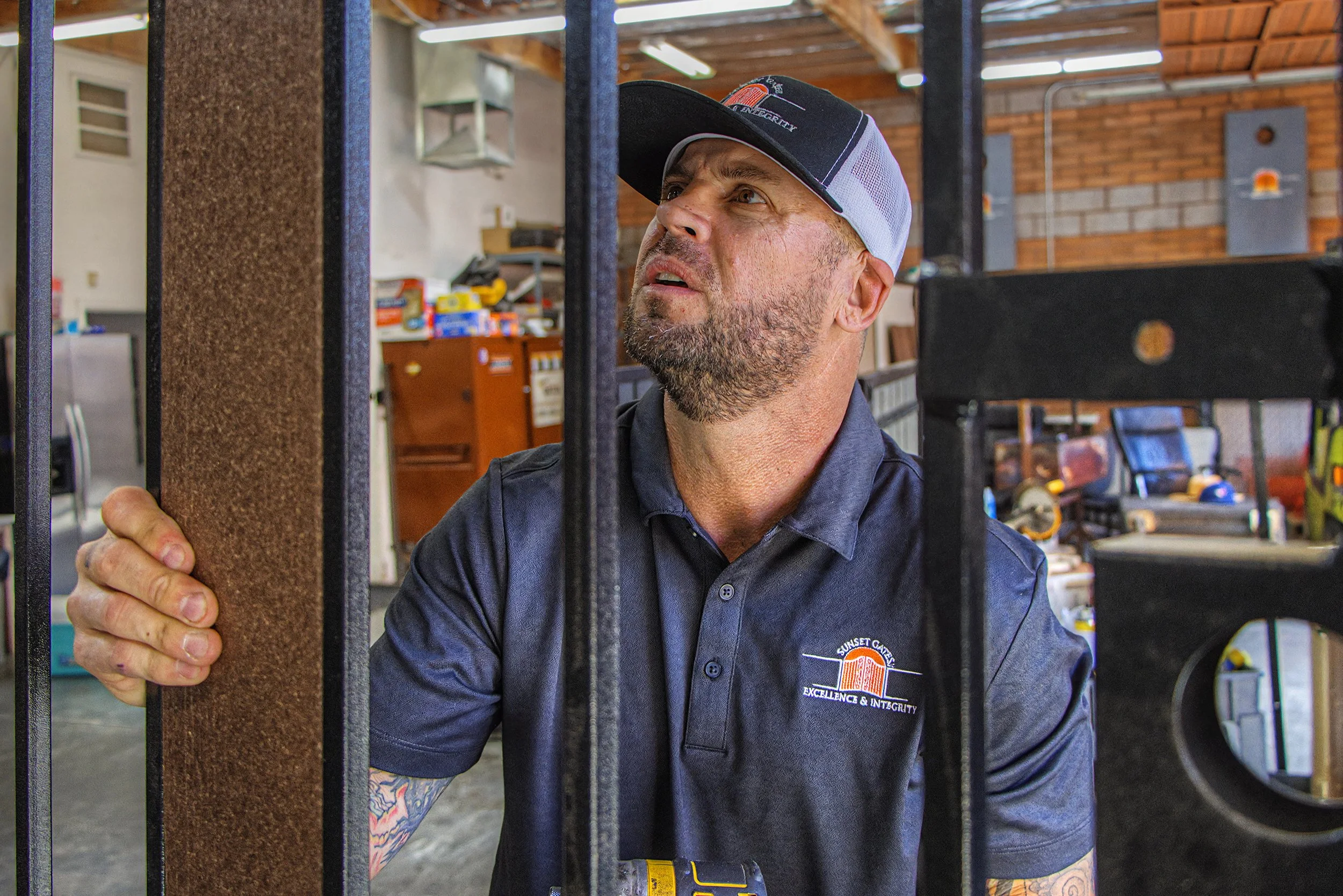 A man with a beard and tattoos, wearing a black shirt and a baseball cap, is looking through a metal fence in a workshop or garage setting. Shelves with tools and supplies are visible in the background.