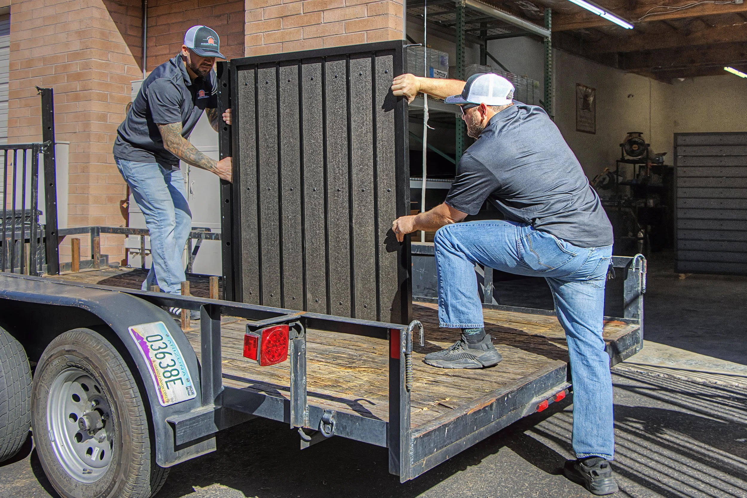 Two men are loading or unloading a large black object onto a flatbed trailer. One man is kneeling, and the other is standing, both wearing gray shirts, jeans, and baseball caps. They are working in an outdoor area near a brick building and a garage.