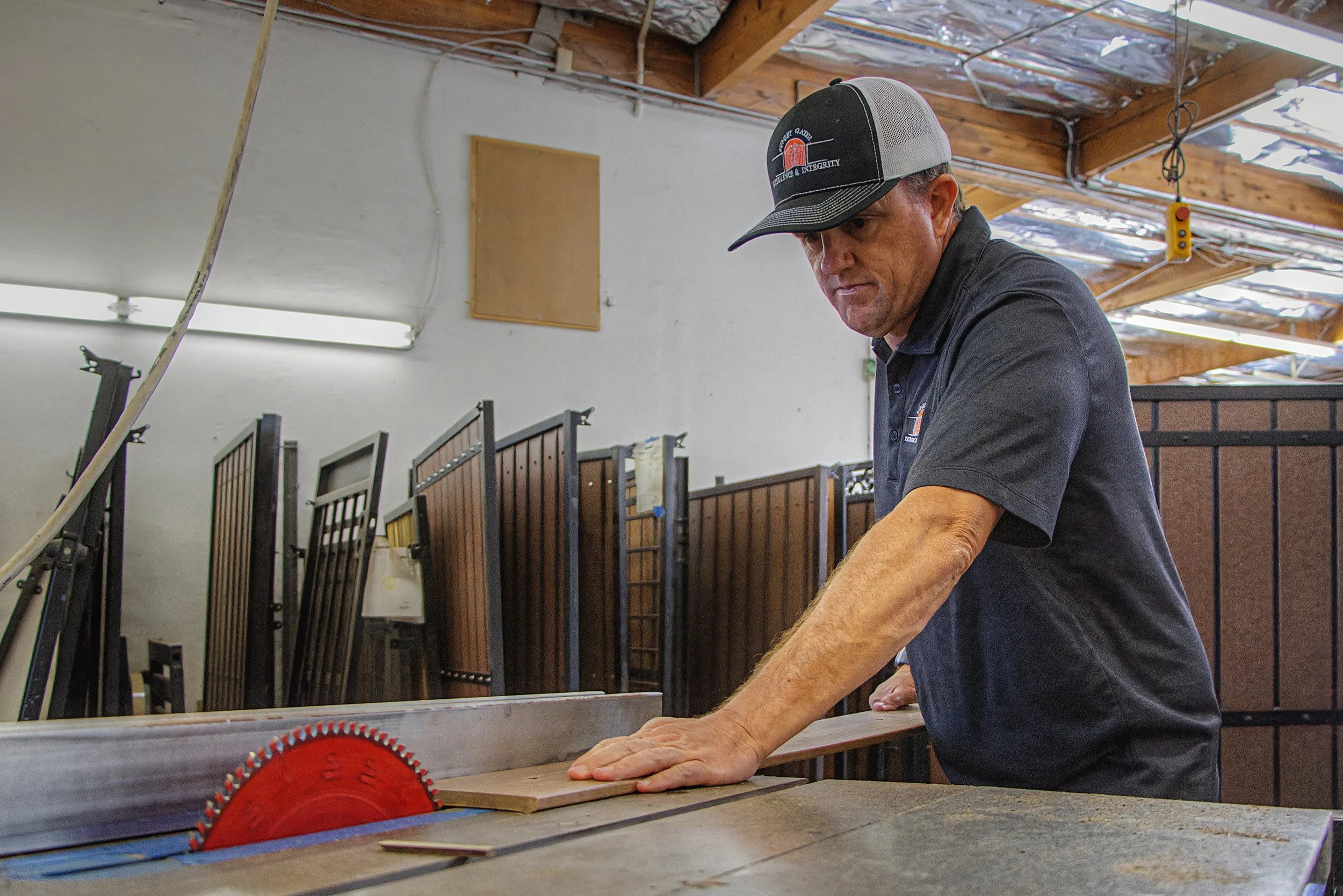 A man in a black cap and shirt is working with his hands on a piece of wood at a workbench in a woodworking shop. The shop has a wooden ceiling with exposed beams and various woodworking tools and materials in the background.