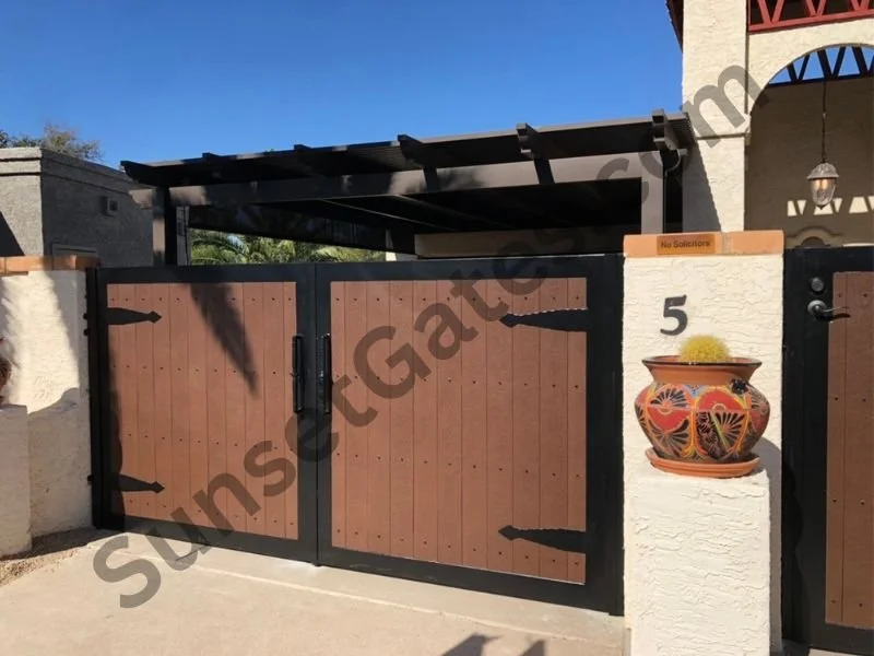 A wooden gate with black accents, part of a residential property with a white stucco wall, potted plant, house number 5, and blue sky.