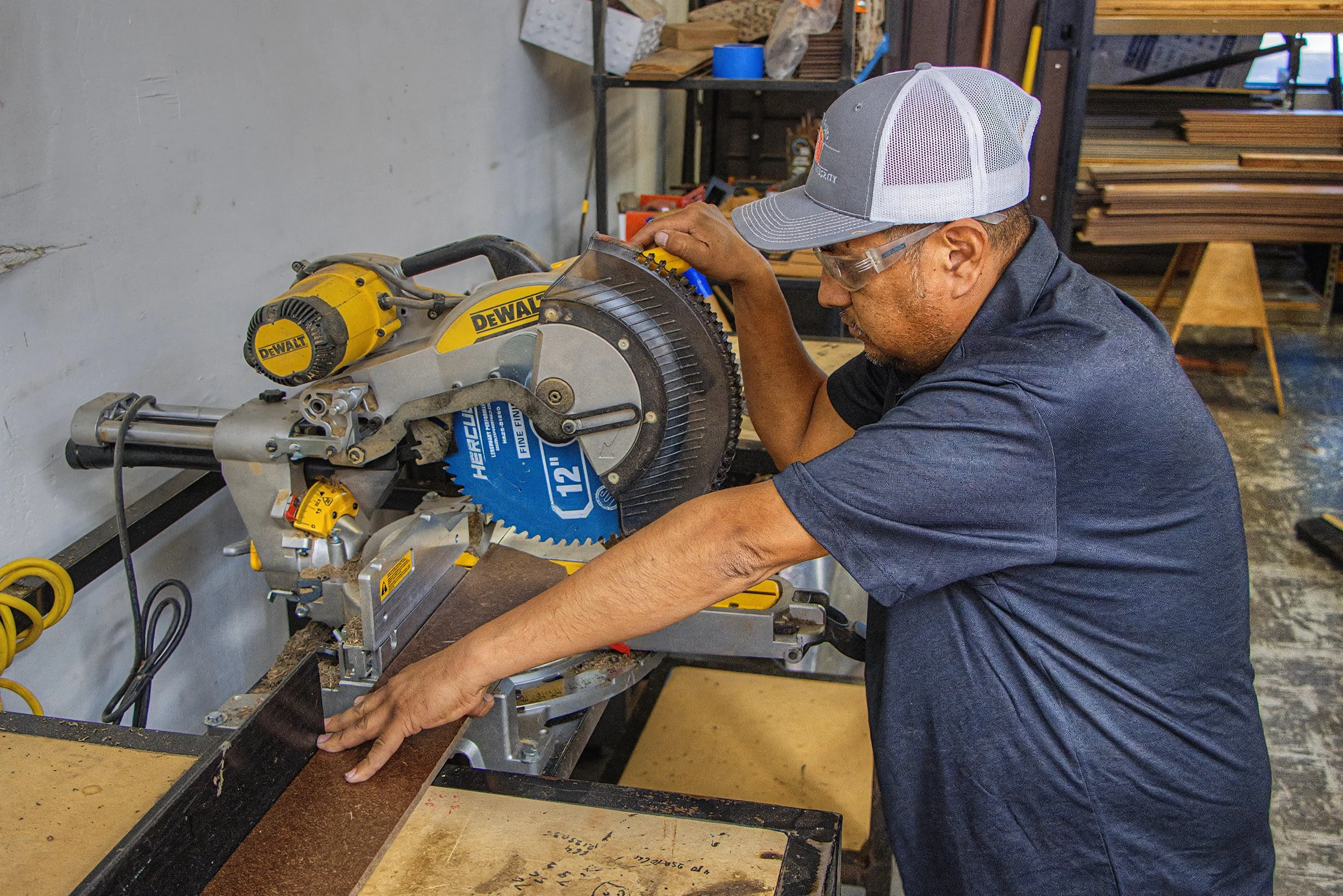 A man wearing safety glasses and a baseball cap operates a large yellow DeWalt miter saw, cutting a piece of wood in a woodworking workshop.