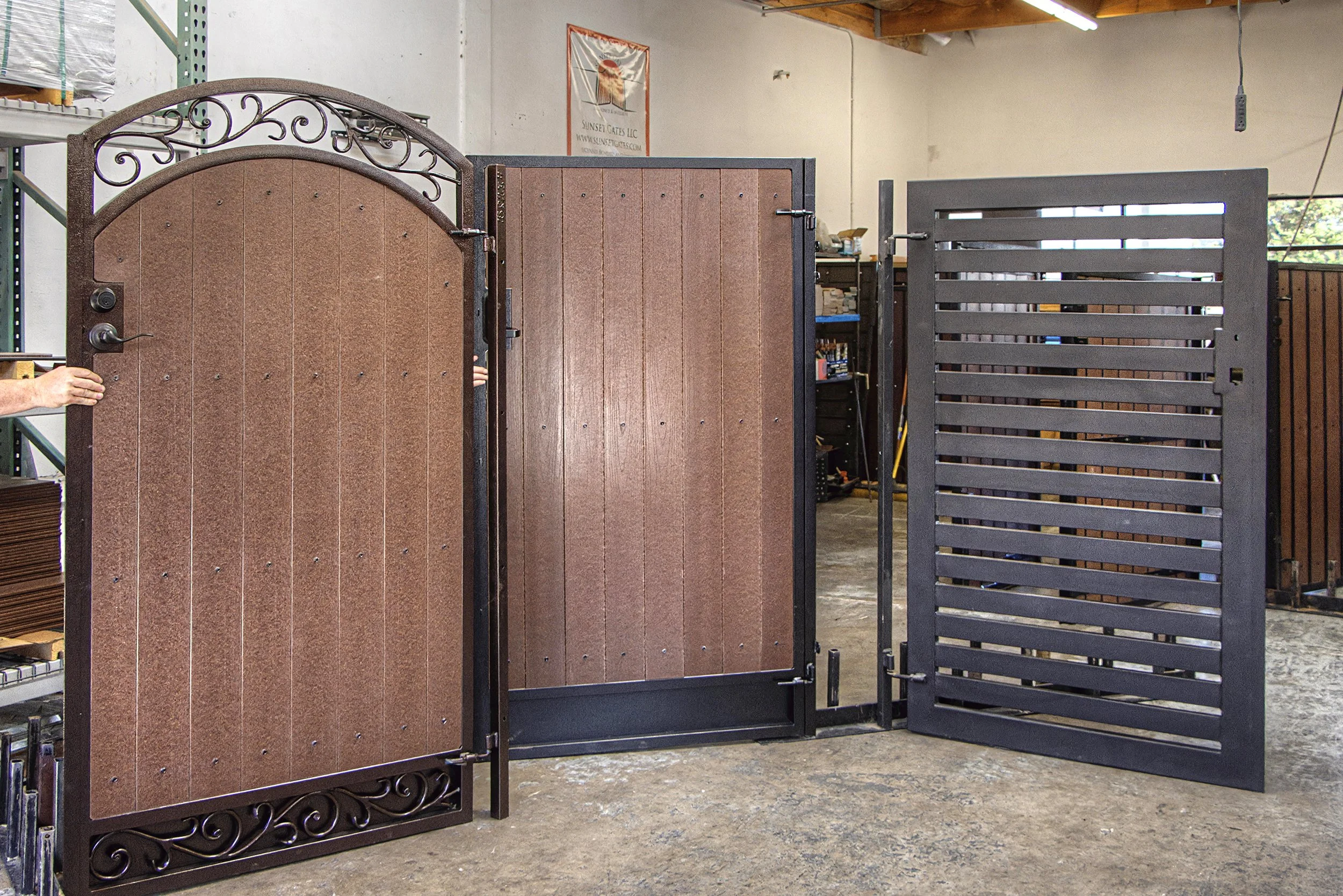 A brown decorative metal gate with a curved top and ornate scrollwork, standing open in a workshop with shelves and other gates around.