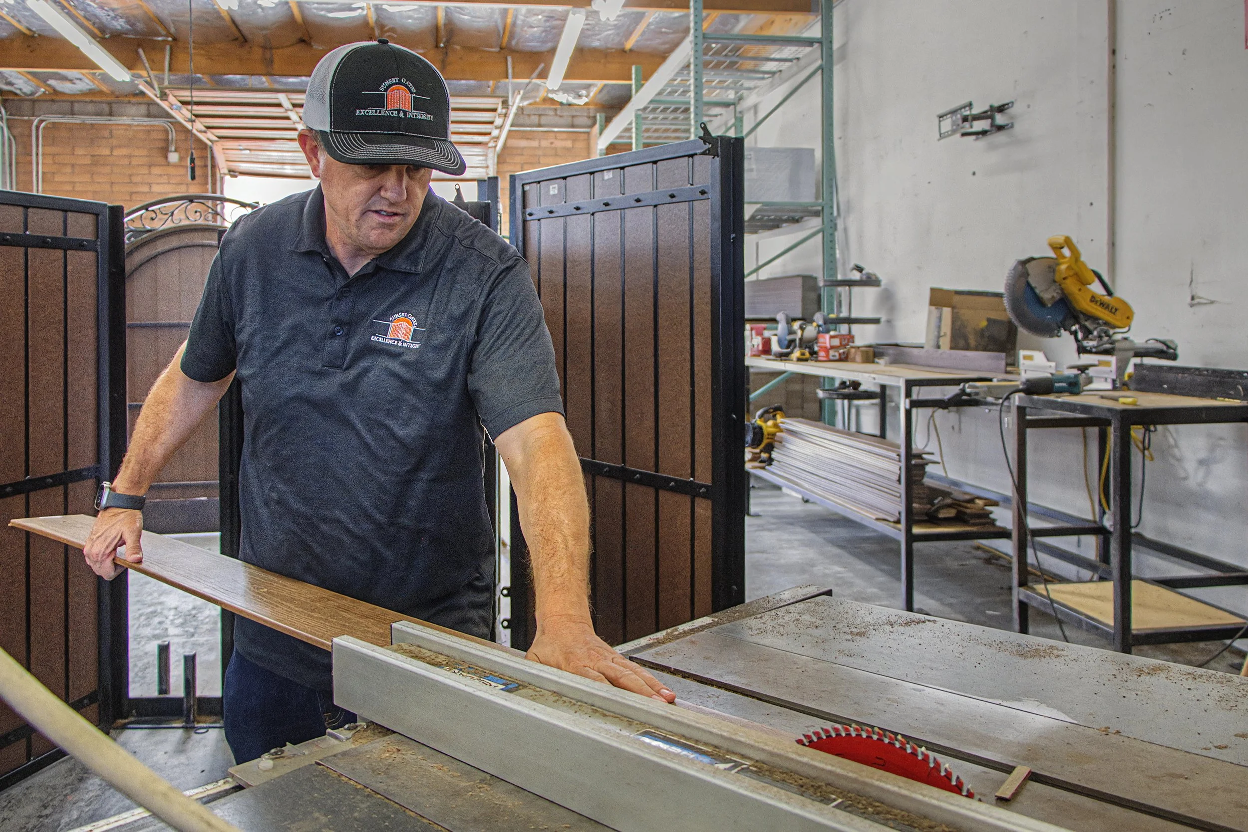 A man wearing a black baseball cap and matching black polo shirt with a logo, working on a woodworking or carpentry project in a workshop. He is holding a piece of wood and standing by a worktable with various tools around.