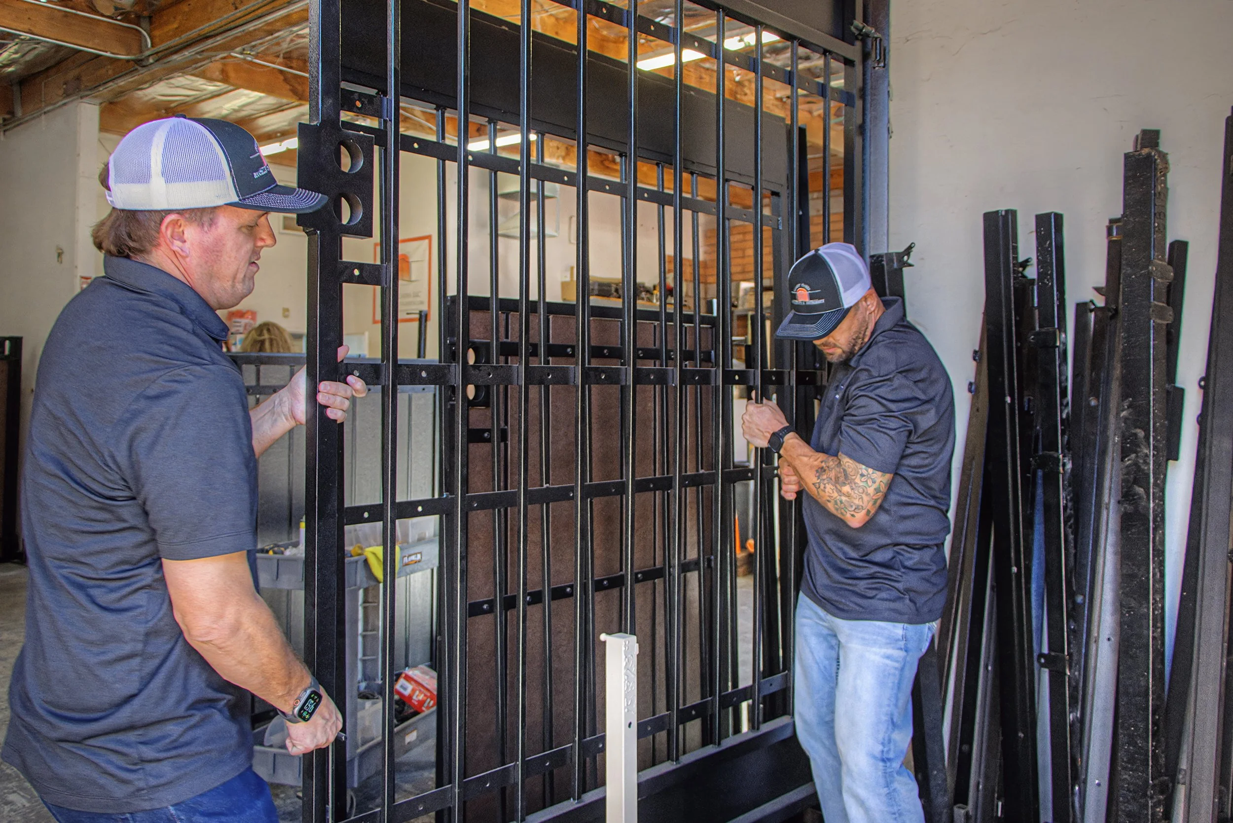 Two men are assembling a large black metal cage or enclosure inside a warehouse or workshop. One man is wearing a navy polo shirt and a trucker hat, while the other is wearing a black jacket and a baseball cap. They appear to be working together to secure or adjust the metal bars.