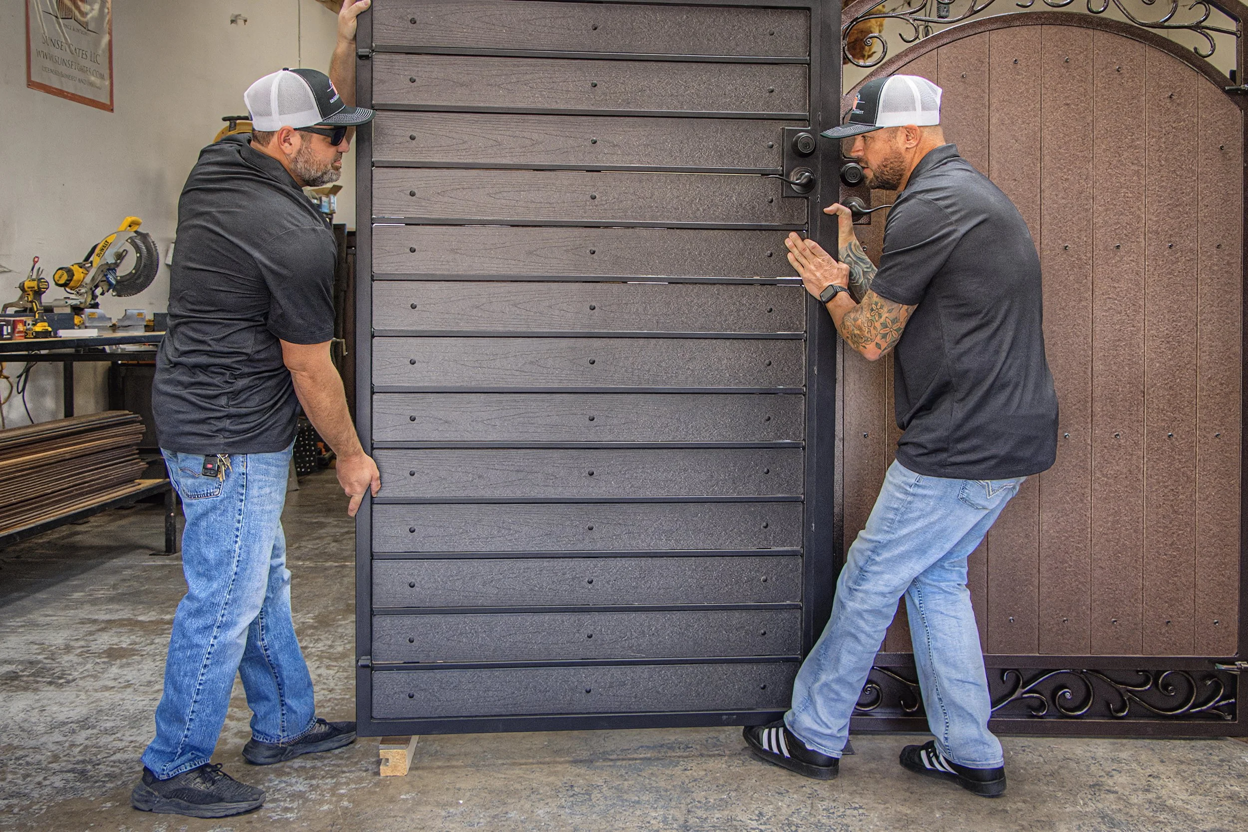 Two men are lifting and positioning a large, dark-colored, wooden, multi-drawer cabinet or storage unit in a workshop or garage setting. They are wearing casual work clothes, including baseball caps and sunglasses.