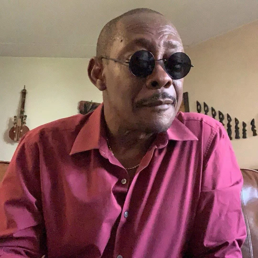 Man wearing round black sunglasses and a pink button-up shirt, sitting on a couch indoors.