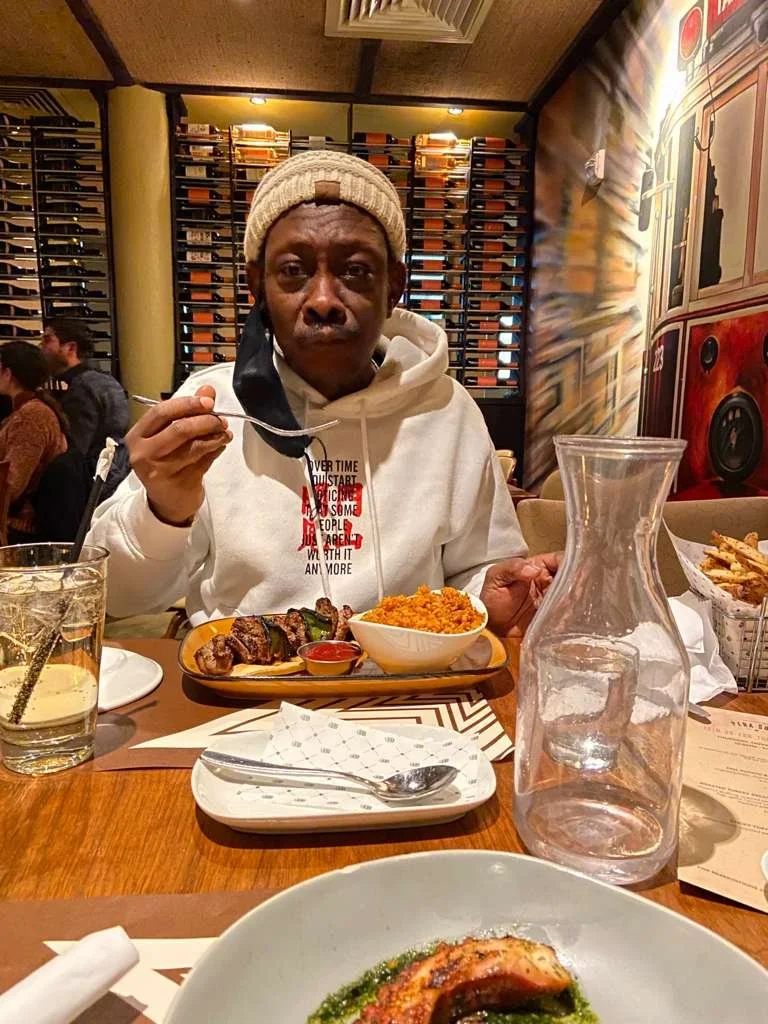 A man in a white hoodie and beige knit cap sitting at a restaurant table, holding a fork. The table has dishes including grilled meat, rice, water, and a glass pitcher. The background features shelves with wine bottles and a colorful mural.