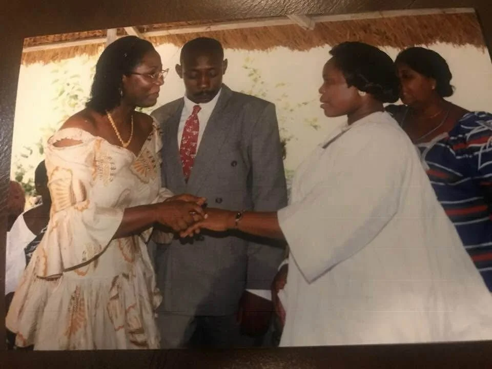 A woman in a patterned dress holding hands with another woman in a white dress, engaging in a handshake or exchange, with a man in a suit and two women in the background observing.