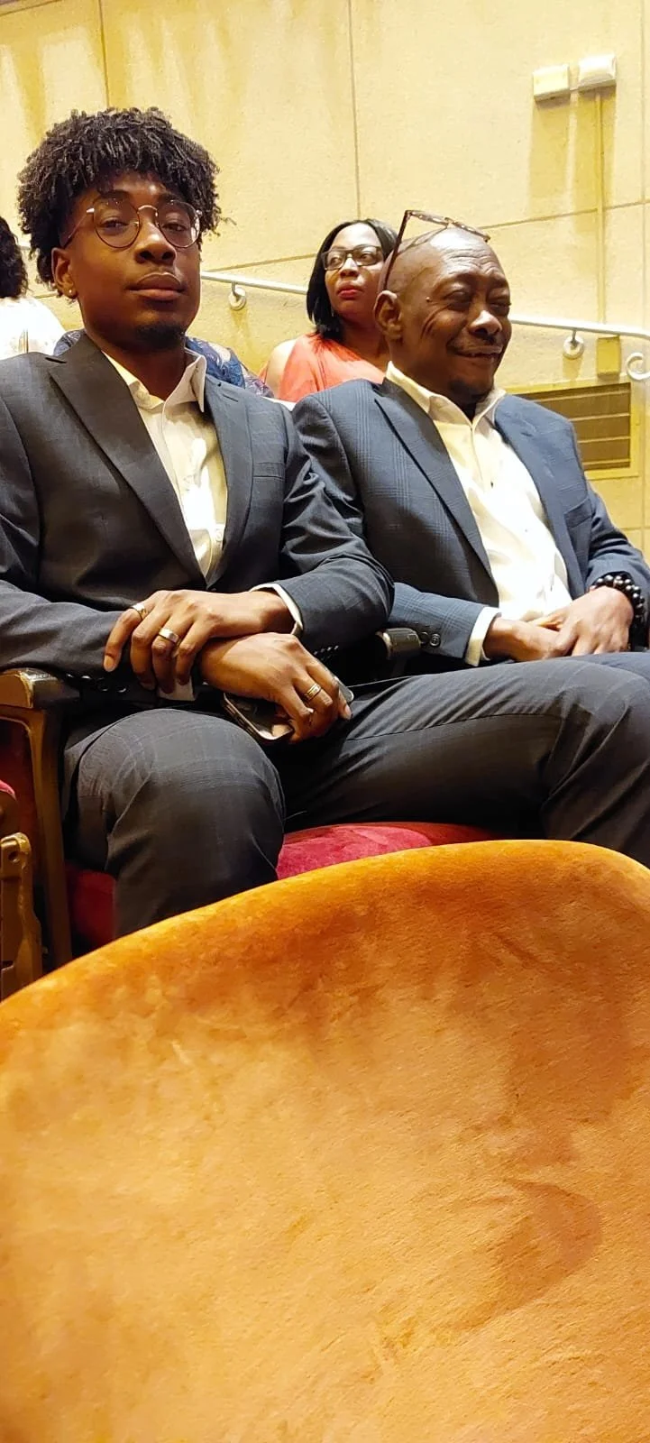 Three people seated in a conference or auditorium, with two men in the foreground wearing suits and a woman in the background wearing glasses and an orange top.