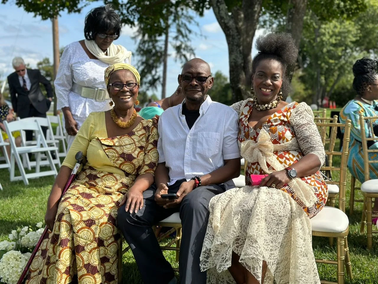 Three people sitting outdoors at a sunny event, smiling and dressed in colorful traditional African attire. A woman on the left has a cane, and a woman on the right holds a pink item.