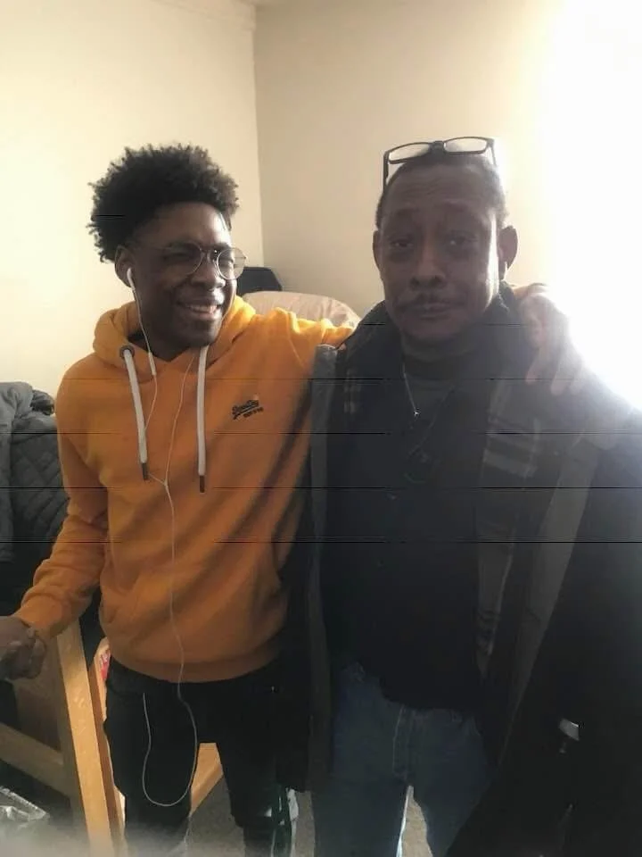 Two men standing close together indoors, one wearing a yellow hoodie and the other wearing a dark jacket, with the man on the left smiling and the man on the right looking serious.
