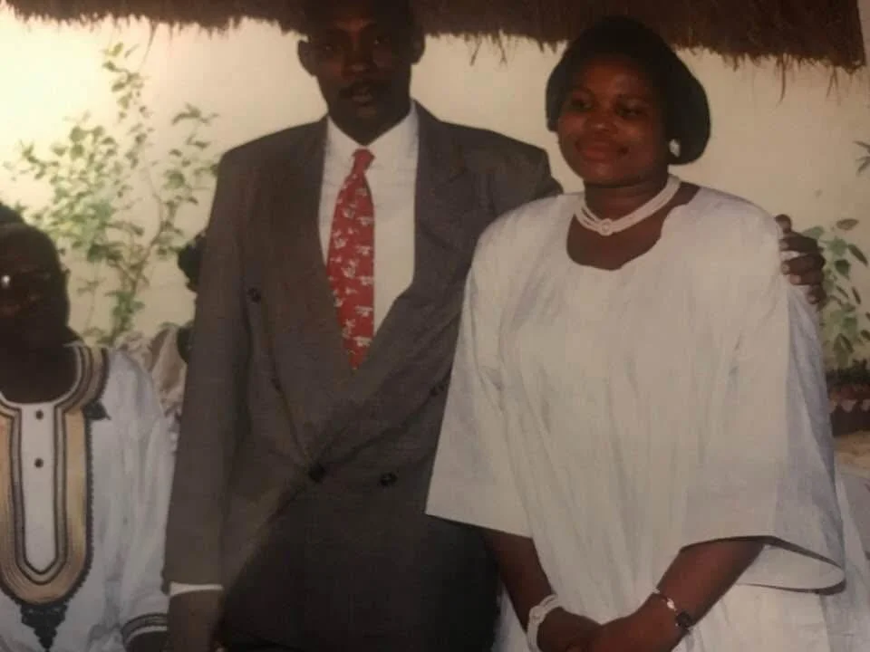 A group of three people, two women and one man, standing together and posing for a photo outdoors. The man is in the middle, wearing a grey suit and a red tie. The woman on the right wears a white traditional dress with jewelry, and the woman on the left wears a white dress with black and gold embroidery.