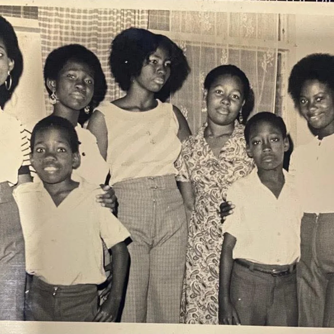 Black and white photograph of a group of seven African American people, including men, women, and children, posing indoors with curtains and a wall in the background.