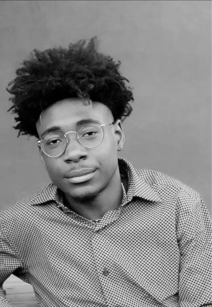 A young man with glasses and textured curly hair wearing a checkered shirt, looking at the camera against a plain background.
