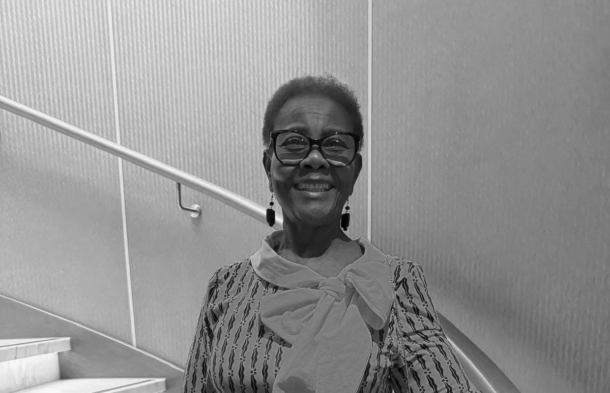 A smiling elderly woman with short hair, wearing glasses, earrings, and a striped blouse with a large bow, standing against a textured wall with stairs and a handrail in the background.