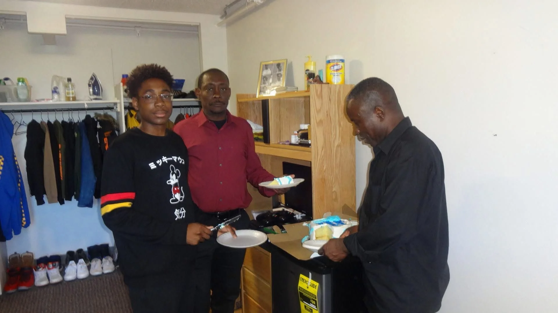 Three men in a room with a bookshelf and clothing rack. Two men hold plates with food, while the third man is preparing food. Shoes are on the floor near the clothing rack.