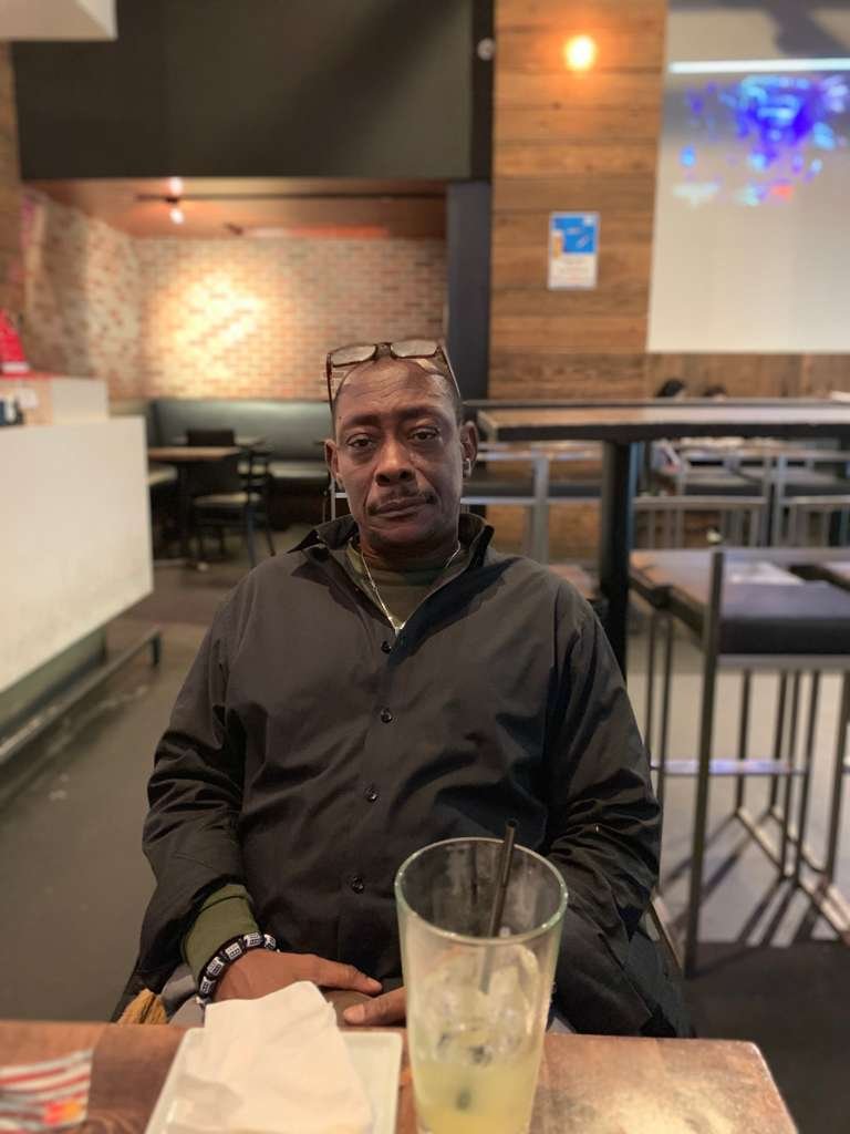A man sitting at a table in a restaurant or bar with a drink and tissue on the table, wearing a black jacket and glasses on his head.