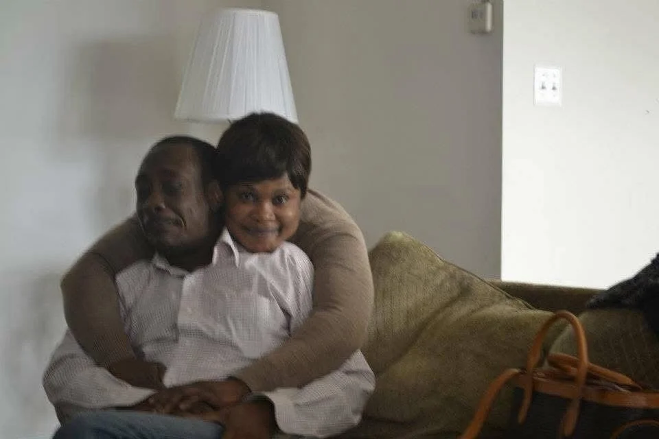 A man and a child sitting on a couch, hugging each other and smiling, in a living room with a lamp and a bag nearby.