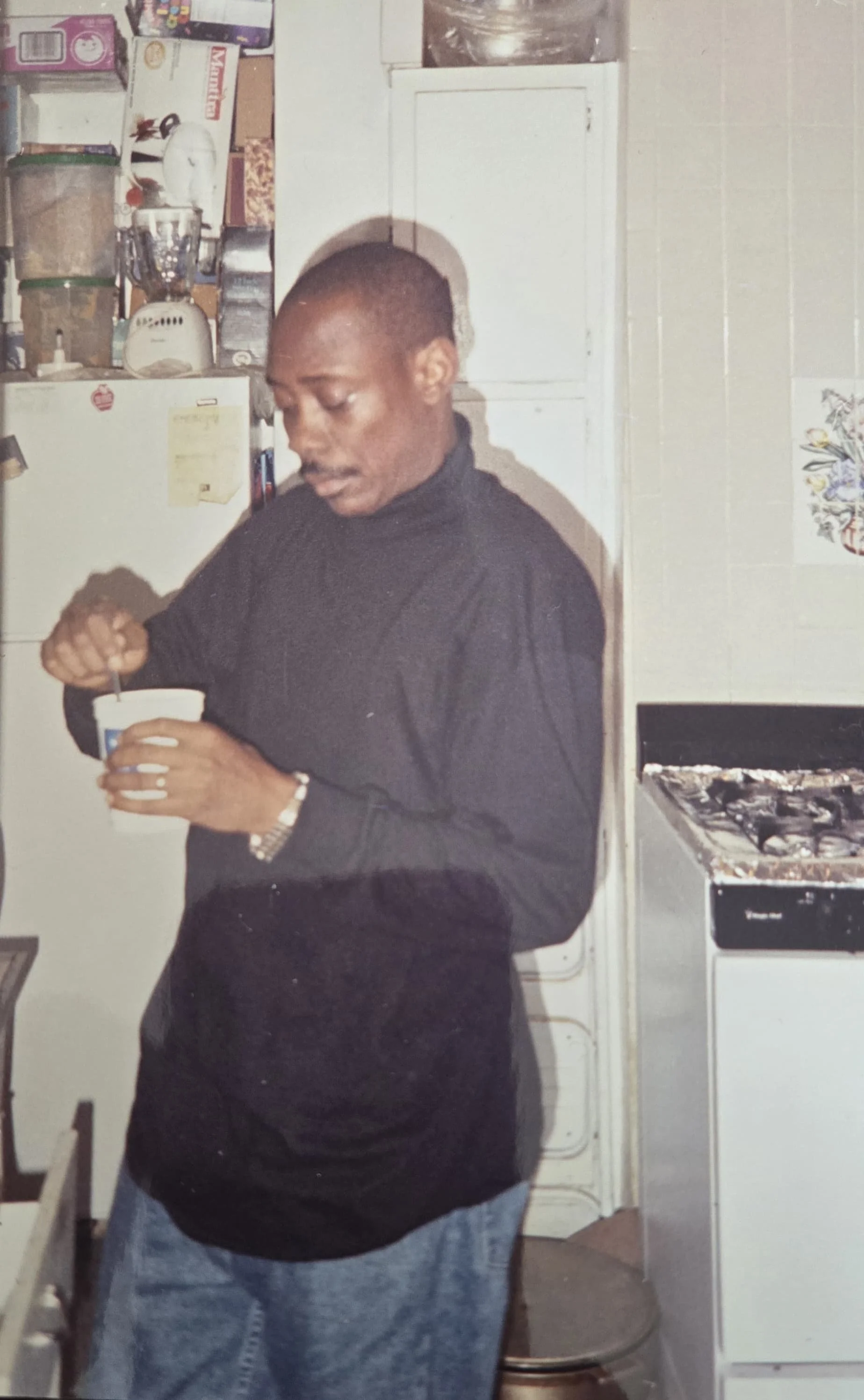 A man in a black sweater and gray jeans standing in a kitchen, stirring something in a cup.