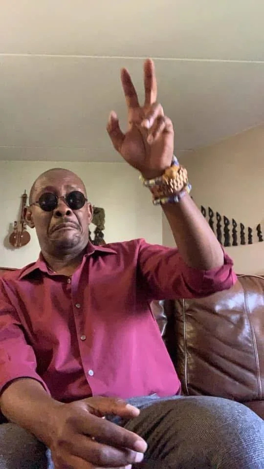 A man sitting on a brown leather couch, wearing a maroon shirt and sunglasses, making a peace sign with his right hand. He has bracelets on his wrist.