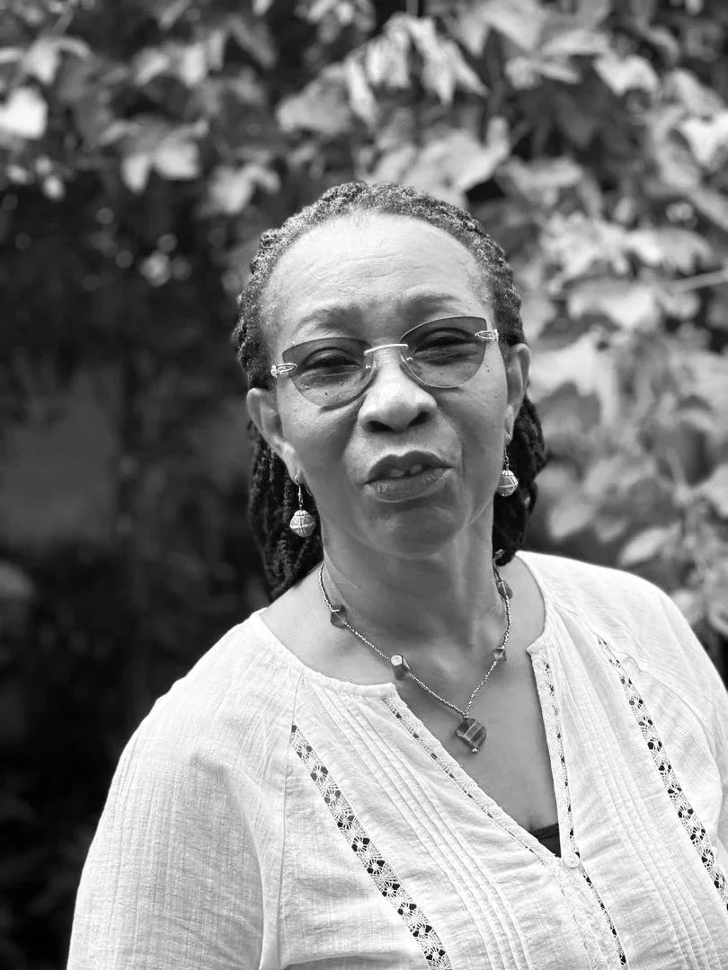 A black and white portrait of a middle-aged woman with glasses, earrings, and a necklace, standing outdoors in front of leafy foliage.
