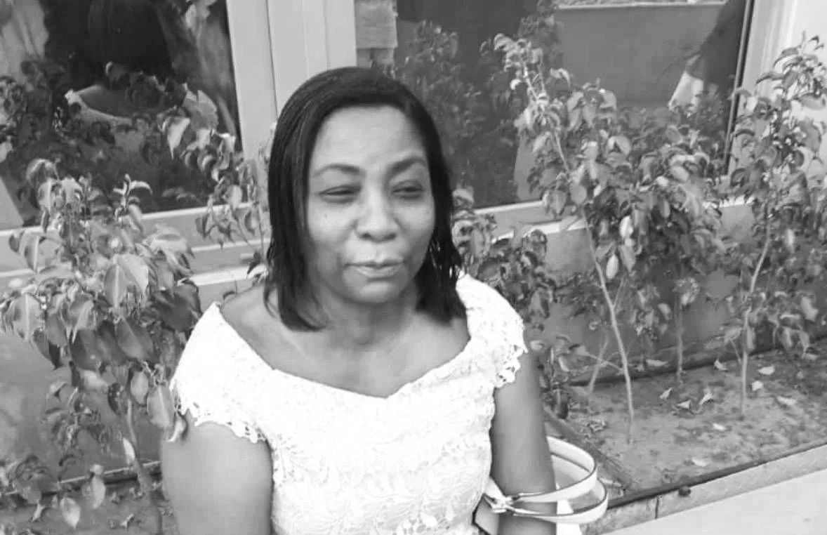 A woman with shoulder-length dark hair sitting outside next to plants, wearing a short-sleeved lace top, with her eyes partially closed.