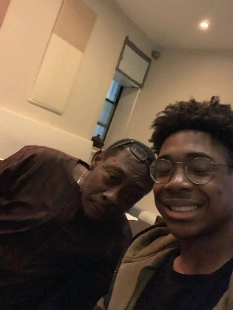 Two men taking a selfie inside a room. One is an older man with glasses and a brown shirt, the other is a younger man with glasses, smiling and wearing a hoodie.