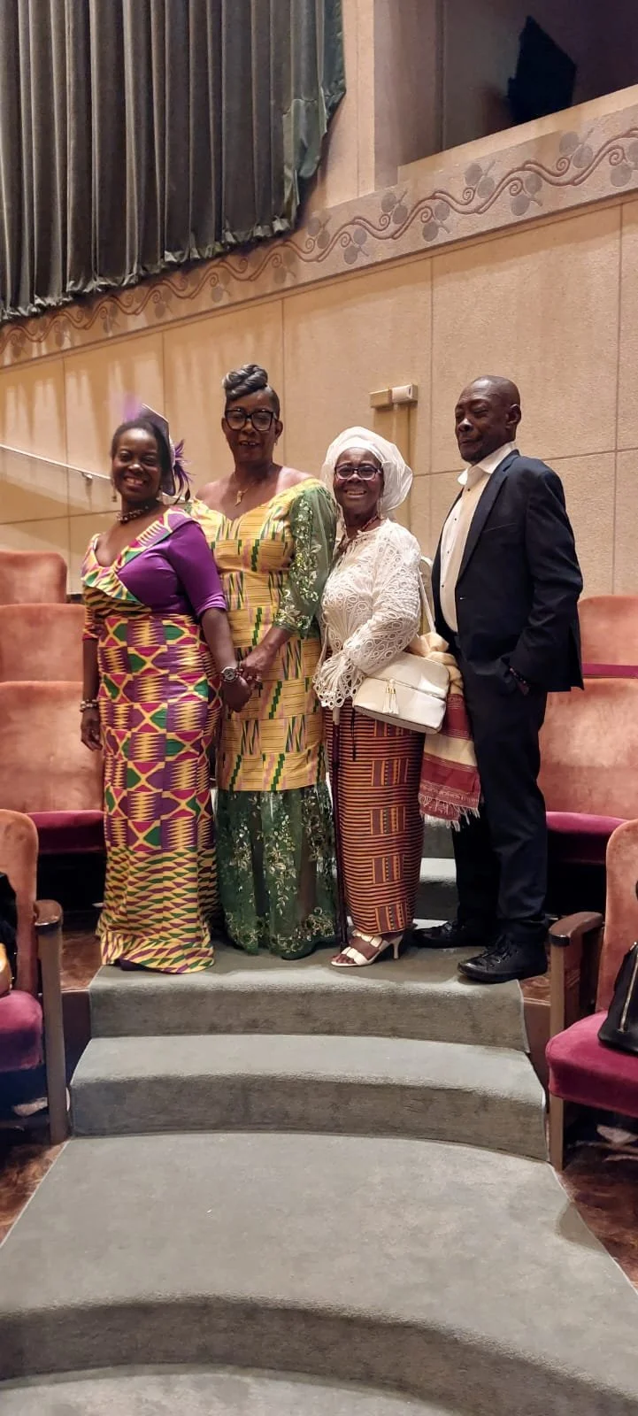 Four individuals dressed in colorful traditional and formal attire pose for a photo in an indoor venue with beige walls and orange seating.