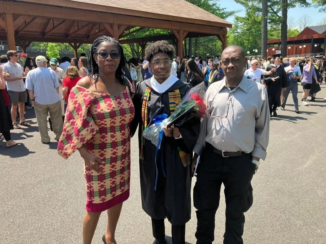 A graduate in a black cap and gown holds a bouquet of flowers, standing with two adults during a graduation ceremony outdoors.