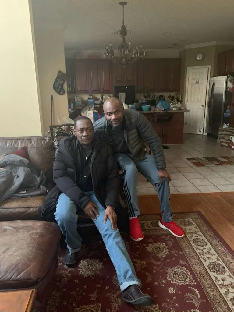 Two men sitting on a brown leather couch in a living room with an open kitchen in the background, one wearing a black jacket and jeans, and the other in a black jacket and red shoes.