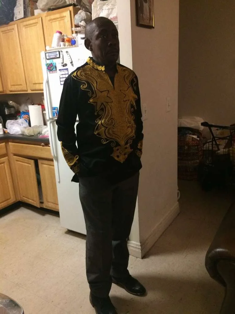 A man standing indoors in a kitchen with wooden cabinets, wearing a black shirt with gold embroidery and dark pants.