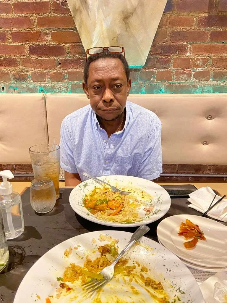 A man sitting at a restaurant table with leftover food on plates, a glass of water, a glass of iced tea, a bottle of hand sanitizer, and napkins, in front of a brick wall background.