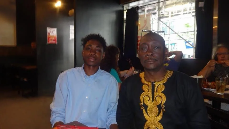 Two men sitting at a restaurant table, the man on the left in a light blue shirt and the man on the right in a black shirt with gold embroidery, with other diners and a window in the background.