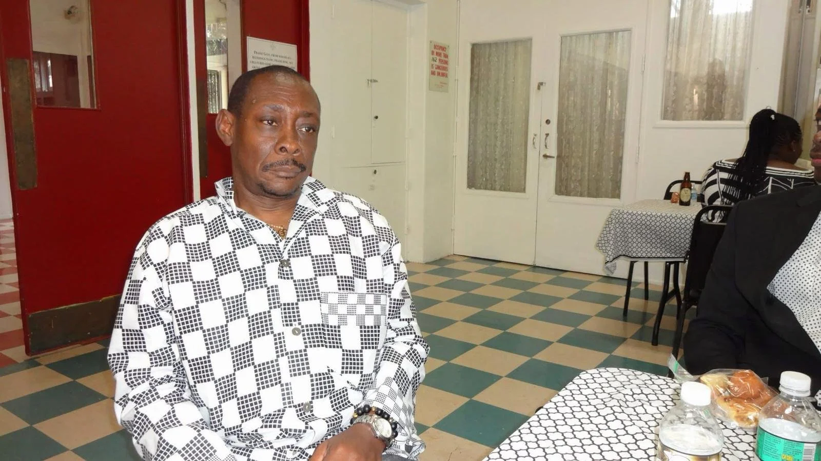 A man with a serious expression sitting at a table in a restaurant, wearing a black and white patterned shirt.