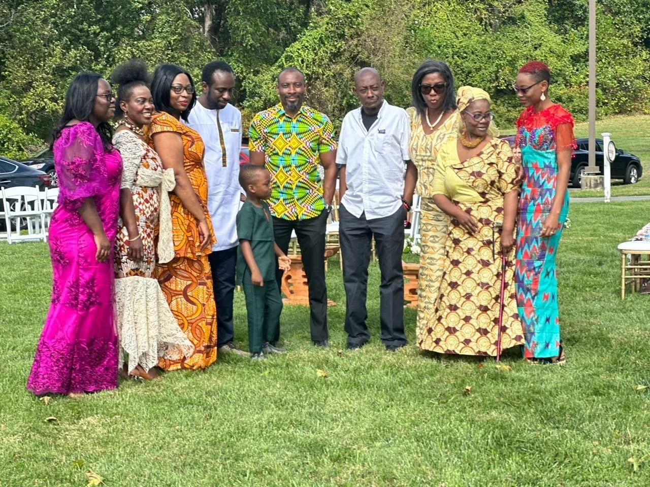 Group of ten people standing outdoors on grass, dressed in colorful traditional and modern clothing, with trees and parked cars in the background.