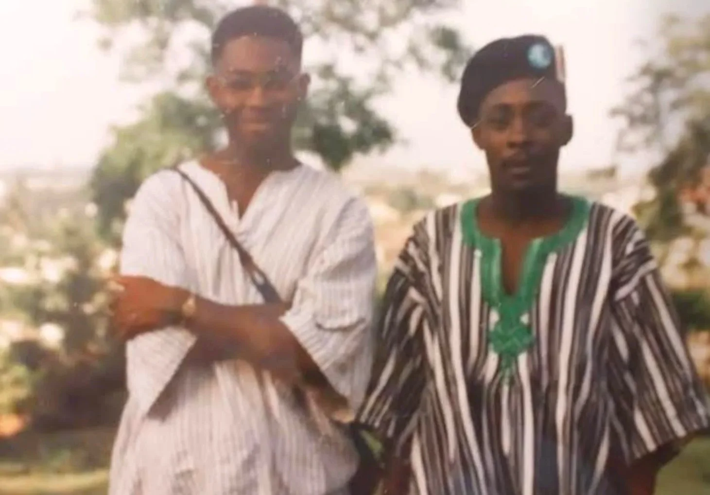Two women standing outdoors, one with glasses and a white and gray striped dress, the other with a headscarf and green top with a black and white striped cape, with trees in the background.