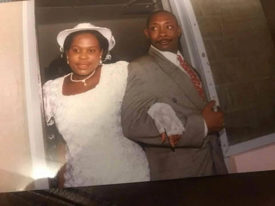 A couple dressed in formal wedding attire, standing close together, with the woman wearing a white wedding dress and hat, and the man in a gray suit with a red tie.