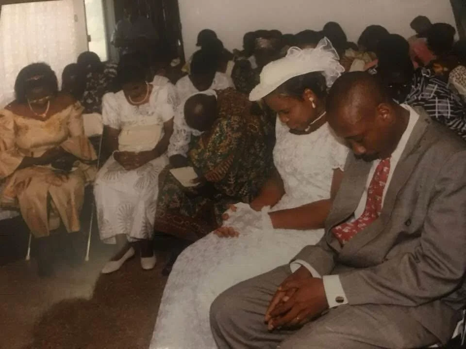 A group of people attending a wedding ceremony, with the bride and groom sitting in front, holding hands and praying.
