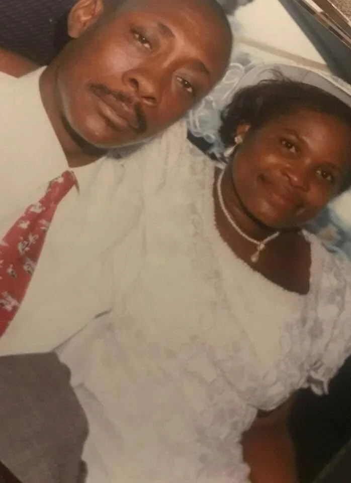 A man and woman taking a selfie together, both dressed in white clothing. The man has a mustache and goatee, and the woman is wearing jewelry and has short hair.