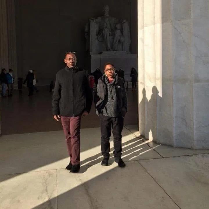 Two young boys standing inside the Lincoln Memorial in Washington, D.C., with a shadow cast on the floor, and the seated statue of Abraham Lincoln visible in the background.
