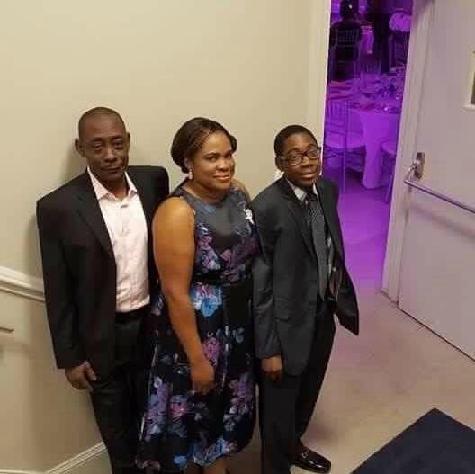 Three people, two men and one woman, dressed in formal attire, standing together indoors near a door and wall.