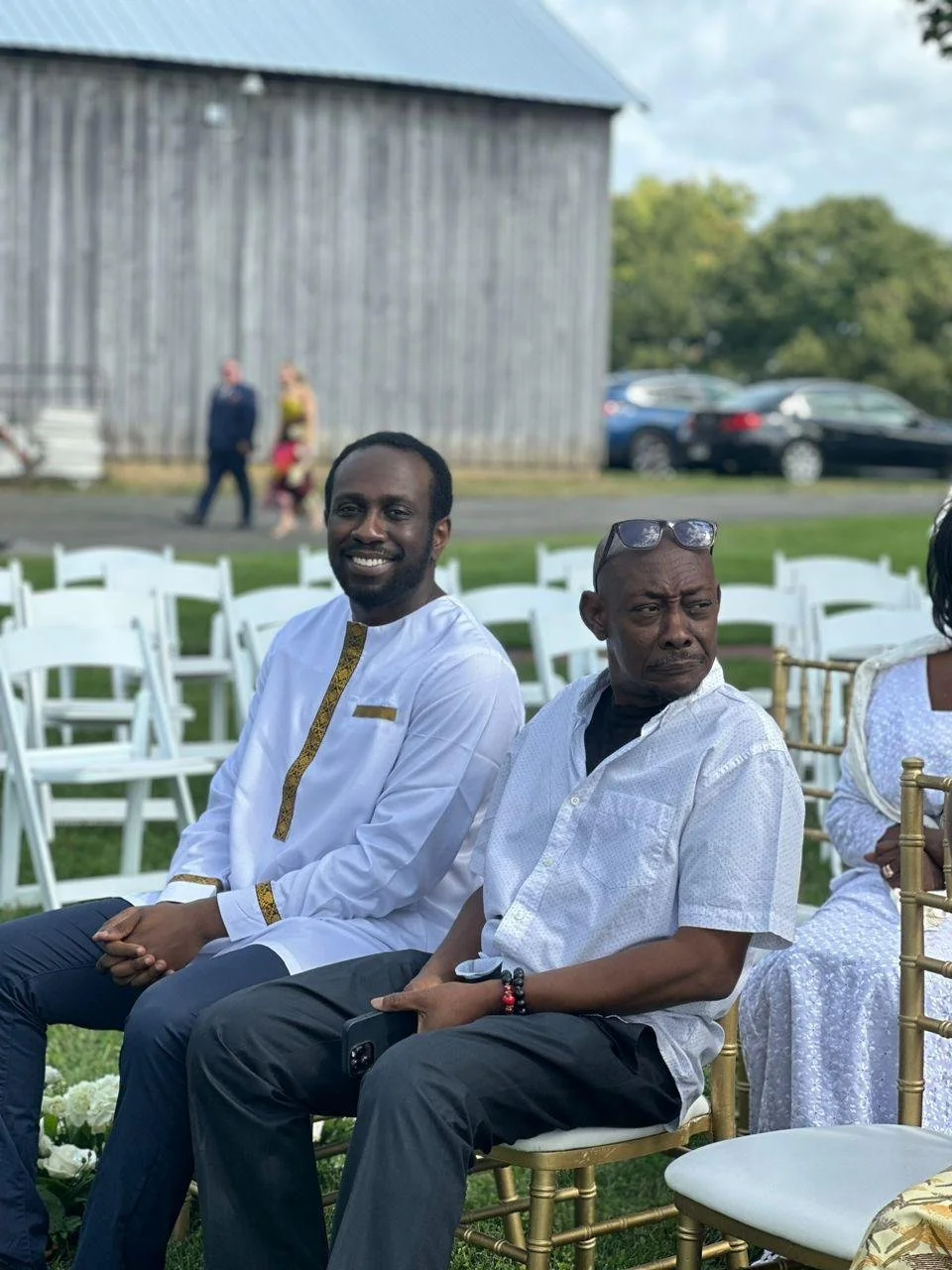 Two men sitting outdoors on white chairs at an event, with a rustic wooden building and several cars visible in the background.