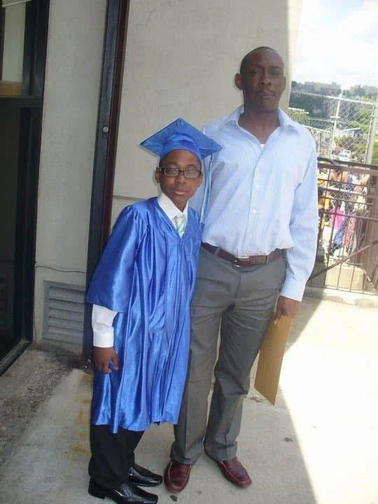 A young boy in a blue graduation gown and cap stands next to an adult man in a white shirt and gray pants, on an outdoor balcony, celebrating a graduation.